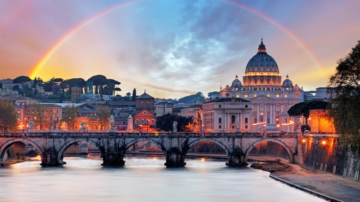 An ancient bridge going across the river in Rome with a rainbow in the city skyline.