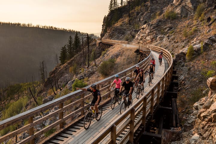 A group of people riding bicycles across a bridge that runs across the side of a mountain.