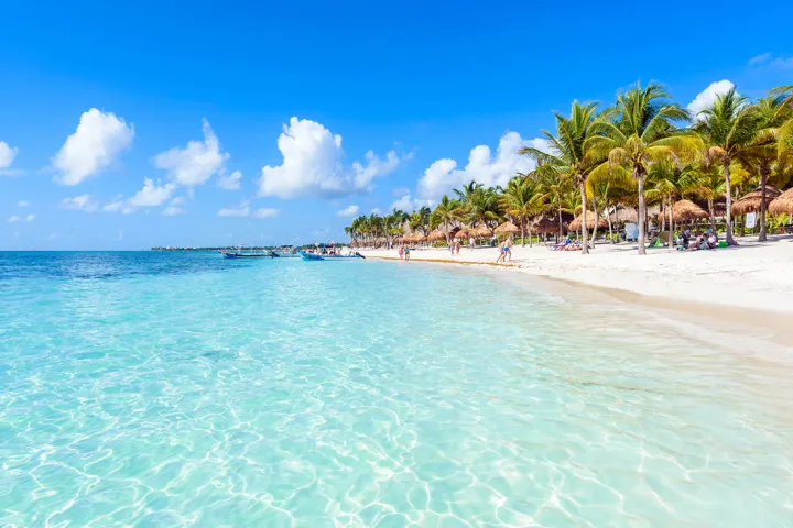 Shallow clear waters next to a white sandy beach in Tulum, with palm trees lining the back of the beach.