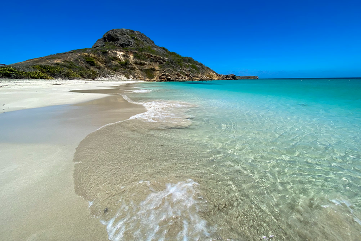 Clear turquoise waves gently washing onto the soft white sandy shore of Isla Caja de Muertos (Coffin Island), with a rocky, green-covered hill rising in the background under a vivid blue sky.