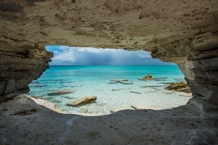 View from inside a rocky cave at Water Cay in Turks and Caicos, overlooking clear turquoise waters, scattered rocks in the shallow sea, and a dramatic sky with distant rain clouds.
