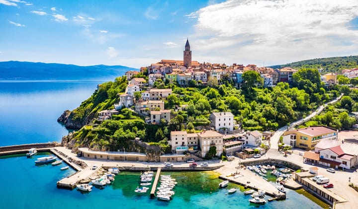 Aerial view of the coastal town of Vrbnik, Croatia, with tightly clustered stone buildings and a prominent church tower perched on a green hillside.