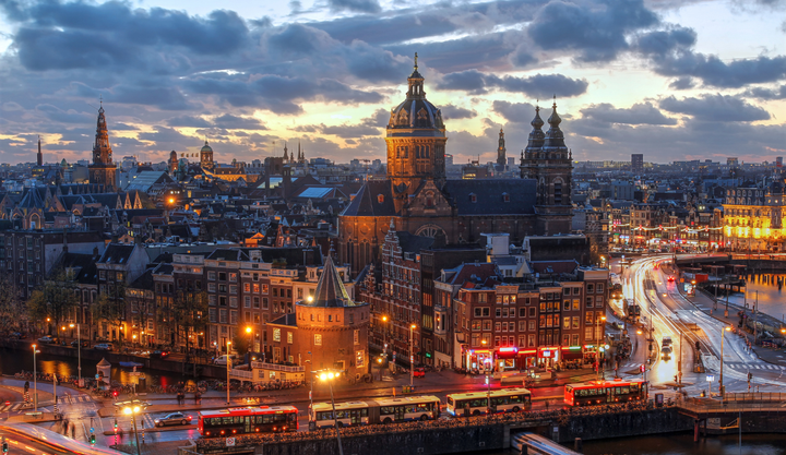 A scenic evening view of central Amsterdam, featuring the Basilica of Saint Nicholas prominently in the background, historic canal houses in the foreground.
