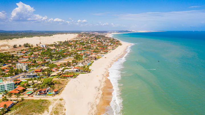 Aerial view of Cumbuco Beach in Ceará, Brazil, showing a long stretch of sandy shoreline bordered by turquoise ocean and colorful coastal homes.