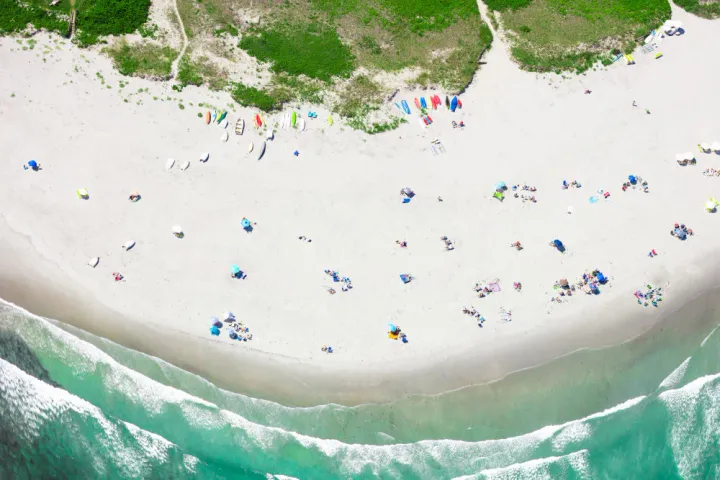 Aerial view of Goose Rocks Beach in Maine showing soft white sand, turquoise waves, scattered beachgoers, and kayaks and paddleboards lined up near grassy dunes.