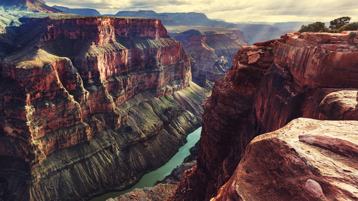 Grand Canyon with dramatic red rock cliffs, steep canyon walls, and the Colorado River winding through the valley floor under a partly cloudy sky in Grand Canyon National Park, Arizona.