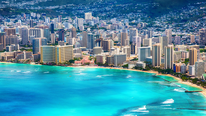 Aerial view of Waikiki Beach in Honolulu, Hawaii, with high-rise resorts lining the turquoise shoreline and lush green hills in the background.