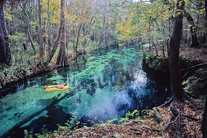 Person floating on a yellow raft in crystal clear waters surrounded by forest.  