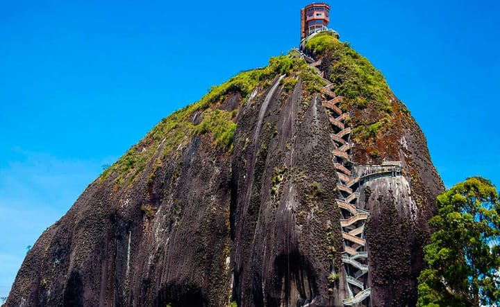 La Piedra del Peñol in Guatapé, Colombia, with a zigzag staircase leading to a viewing tower at the summit, set against a clear blue sky.