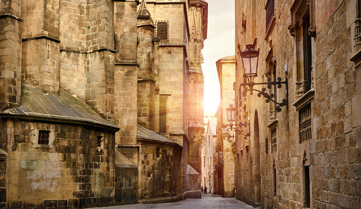 Narrow cobblestone alley lined with medieval stone buildings in Barcelona’s Gothic Quarter, illuminated by warm sunlight and antique street lamps.