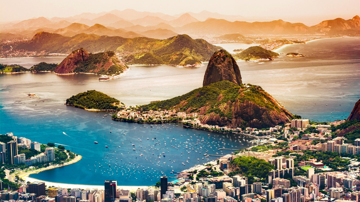 Aerial view of Rio de Janeiro, Brazil, showing Sugarloaf Mountain, surrounding green hills, city buildings, and boats in the blue bay under a hazy sky.