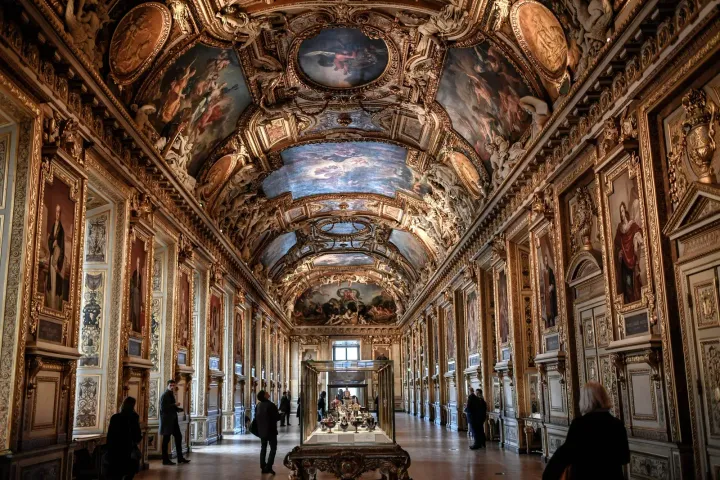 Visitors walk through an ornate gallery in the Louvre Museum, Paris, with gilded walls, framed portraits, and a vaulted ceiling adorned with elaborate paintings.