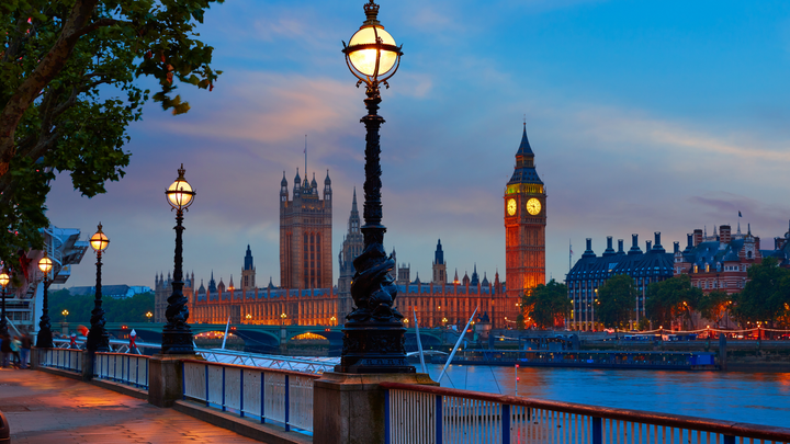 Evening view of Big Ben and the Houses of Parliament in London, seen from the River Thames with street lamps lit and reflections on the water.