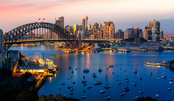 Sailboats anchored in Sidney Harbor at sunset with the city skyline and bridge seen behind them.