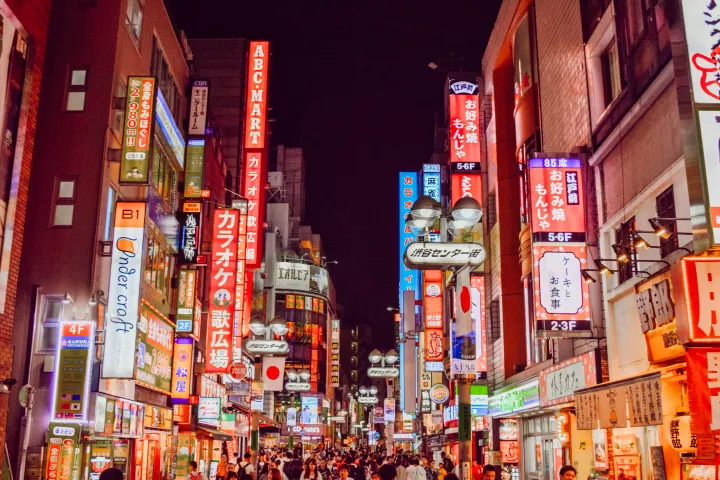 People waling a street with bright lights and street signs in Tokyo at night.