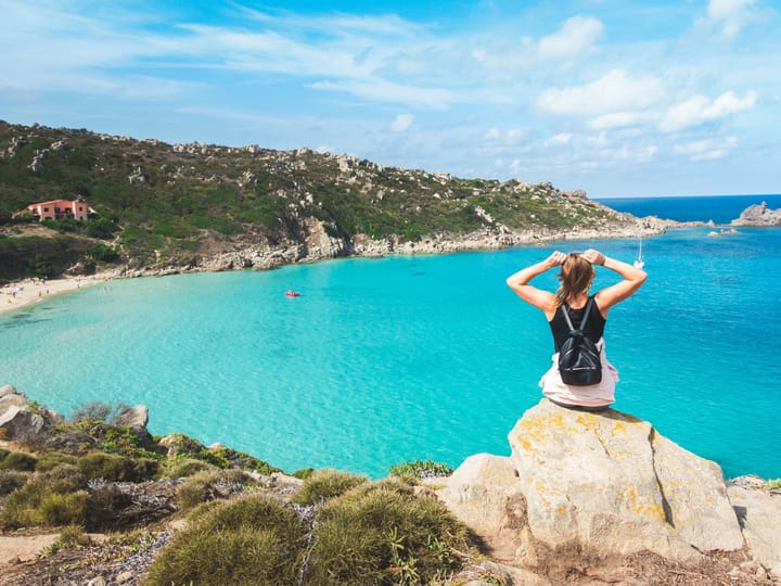 Woman with a backpack sitting on a rock overlooking a beautiful view of the ocean in Sardinia, Italy.
